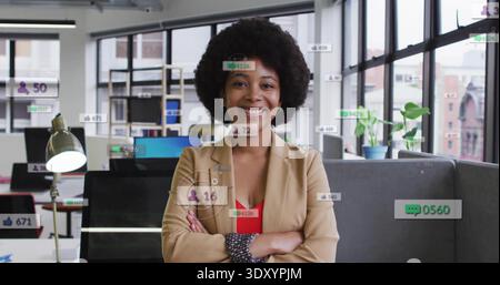 Standing office worker wearing beige blazer, red top, crossing arms in open-plan office, overlays Stock Photo