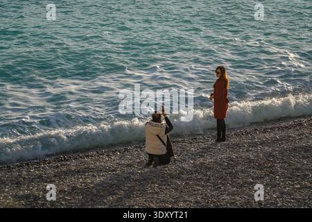 Couple taking pictures along Promenade des Anglais pebble beach shoreline. Turquoise water crashing. Stock Photo
