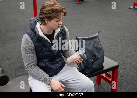 Young man holding protein shaker and sits on gym bench. Training Outdoors. Relaxed and enjoy after workout. Stock Photo