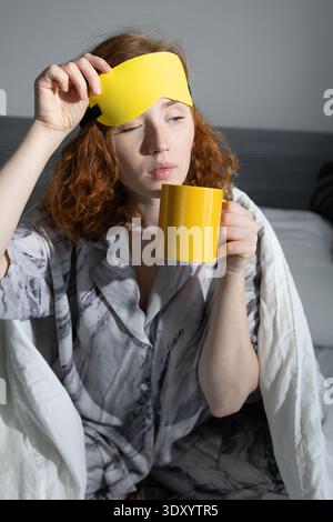 A woman is enjoying a peaceful and cozy morning while sipping coffee, wearing a soft sleep mask and comfy pajamas Stock Photo