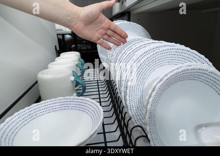 A hand reaches toward neatly arranged clean dishes and mugs in a drying rack. The kitchen setting shows various white plates and cups, all prepared for use. Stock Photo