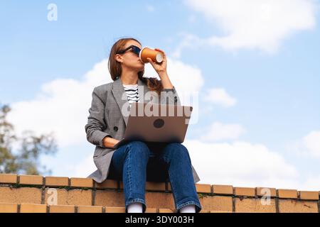Young professional drinking coffee while working remotely on city steps. Stock Photo