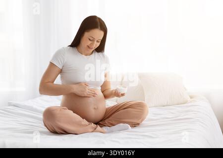 Pregnant young woman gently applying body cream on belly at home Stock Photo