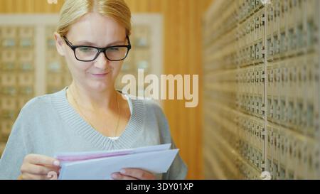 Woman with glasses reading letters near apartment mailboxes, resident checking postal correspondence in residential building hallway Stock Photo