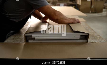 Factory worker packing flat-pack furniture components into a cardboard box for shipping in an industrial warehouse. Stock Photo
