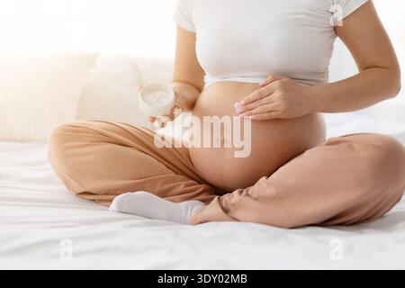 Pregnant young woman applying moisturizing cream on belly while sitting on bed Stock Photo