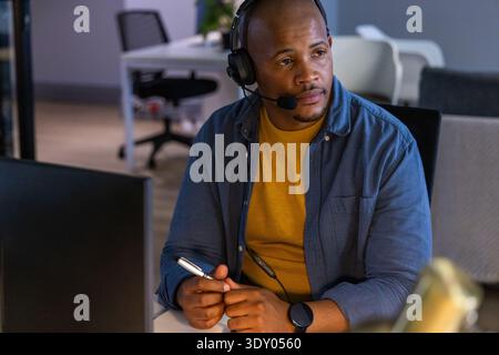 African American man sitting at office desk wearing headset and mustard sweater holding pen Stock Photo