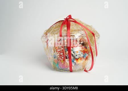 Gift basket wrapped in clear cellophane with red ribbon, filled with assorted chocolates and candy on white background. Stock Photo