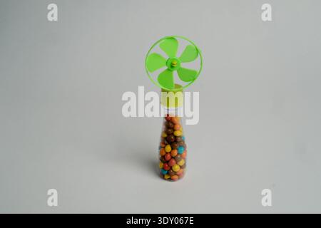 Small handheld candy dispenser with a green pinwheel-style fan top and clear container filled with colorful candy-coated chocolates on a plain light b Stock Photo