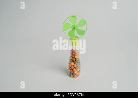 Small handheld plastic candy fan with green propeller top and clear tube filled with colorful chocolate candies, isolated on light gray background wit Stock Photo