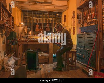 Mature adult luthier meticulously applying varnish to a violin scroll, working on handcrafted string instruments in a cluttered, authentic workshop fi Stock Photo