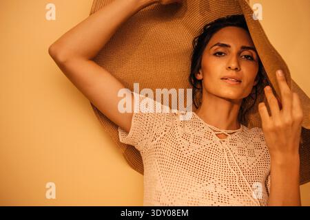 Dark-haired female model in a crochet top and straw hat poses against a warm yellow backdrop, exuding a carefree summer atmosphere and effortless elegance || Model released Stock Photo