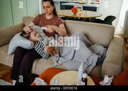 Caring mother measuring temperature of her ill son lying on sofa  Stock Photo
