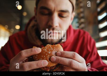 Young man holding and eating a juicy beef burger with bacon on a sesame bun while sitting in a fast food restaurant. Concept of casual dining, quick Stock Photo