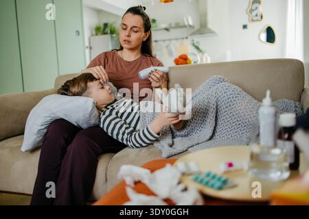 Caring mother measuring temperature of her ill son lying on sofa  Stock Photo