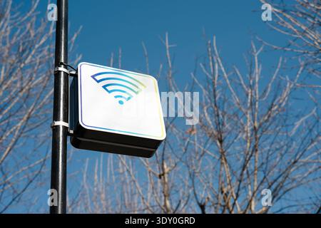 A white square sign with a colorful Wi-Fi symbol mounted on a black metal pole outdoors. The sign indicates a public wireless internet hotspot. Stock Photo