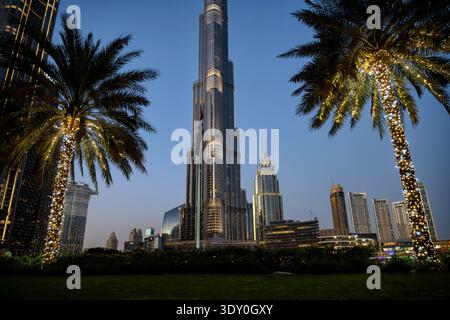 A low-angle view of the Burj Khalifa in Dubai during the blue hour. The iconic skyscraper stands tall in the center, flanked by two palm trees wrapped Stock Photo