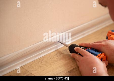 Man fastening plastic floor skirting board with cordless screwdriver on linoleum flooring during interior renovation and baseboard installation. Stock Photo