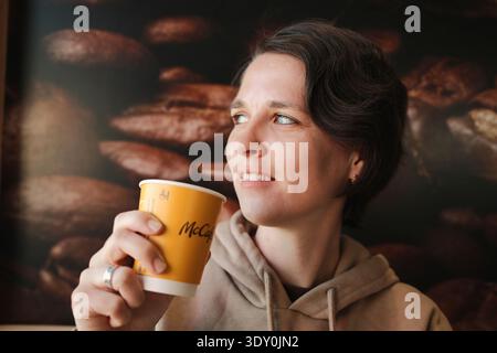 Woman drinking coffee from McCafe paper cup inside McDonalds restaurant interior with coffee beans background. Concept of global coffee chain and Stock Photo