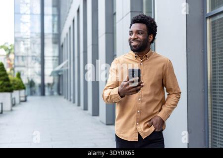 Young man standing outdoors beside a modern office building, holding a mobile phone and looking away with a happy, confident expression, representing success and urban lifestyle Stock Photo