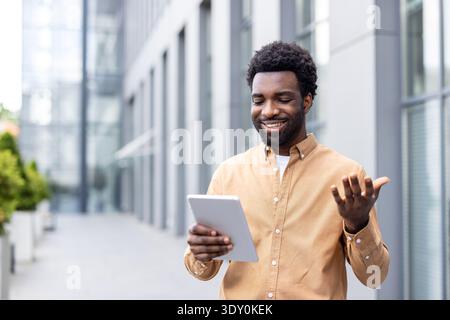 Young man smiling and gesturing while having a video conference on his digital tablet, standing outdoors in a modern urban setting, engaging in remote communication Stock Photo