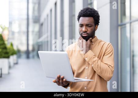 Professional man concentrating on his laptop screen while standing outside a modern building, showing contemplation and problem-solving in a business or academic setting Stock Photo