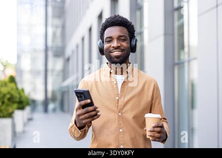 Young man smiling, listening to music on wireless headphones, holding a disposable coffee cup and smartphone while walking outdoors in a modern urban environment Stock Photo