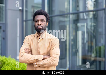 Young man with a serious expression and crossed arms stands confidently in front of a modern glass building, portraying professionalism, determination, and competence in an urban setting Stock Photo