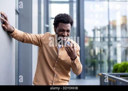 Young black man clutching his chest and using an emergency inhaler against a wall while struggling to breathe during an asthma attack outdoors, urgent relief needed Stock Photo