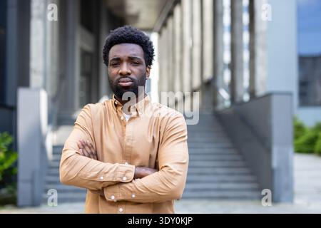 Young man with afro hair standing confidently with crossed arms, looking determined towards the camera against an urban corporate building backdrop, symbolizing strength and professionalism Stock Photo