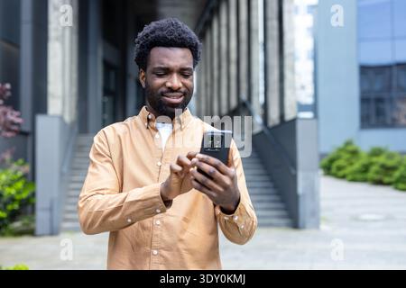 Young adult man smiling while using a smartphone for communication and social media, engaging with digital technology in a contemporary urban environment Stock Photo
