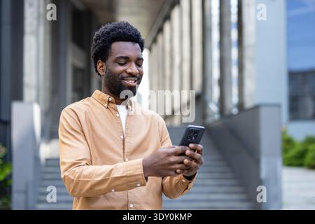 Young african american man standing on an urban street, smiling and interacting with his smartphone, representing communication, social networking, and modern digital lifestyle Stock Photo