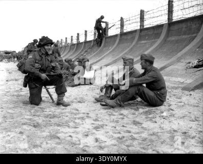 A French Canadian corporal of the Chaudière Regiment of the 3rd Canadian Infantry Division kneeling and speaking to two German prisoners on Juno beach. Other prisoners are sitting a bit further away.  Although the soldier (identified as Victor Deblois) has his Sten gun aiming at the men, the attitude of everyone appears relaxed and Cpl Deblois even appears to be smiling.  On the 6th June 1944 a huge Alllied invasion force involving 7000 ships and 140,000 men successfully landed on the French coast and established five beach-heads. A successful deception campaign meant that much of the German r Stock Photo