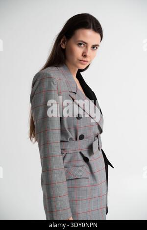 Young brunette woman posing in a studio, wearing an elegant plaid blazer dress with a belted waist, showcasing modern fashion and professional style Stock Photo