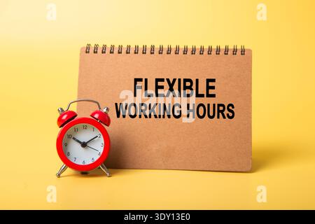 Notepad displaying flexible working hours with a red alarm clock on a yellow background Stock Photo