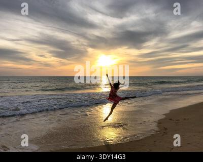 A young woman in a light, flowing summer dress is captured mid-air in a graceful, ballet-inspired jump on a sandy beach at sunset. Stock Photo