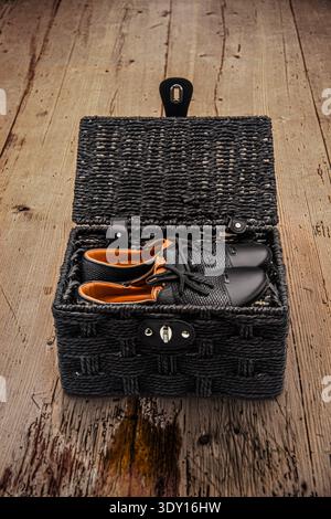 Minimalist still life image of a pair of black laced leather shoes with woven leather in a black wicker basket on a worn ancient wooden table Stock Photo