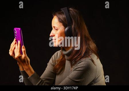 Woman with headphones looking at a smartphone in a dark setting, focused and emotionally engaged with modern technology, mobile communication and digi Stock Photo