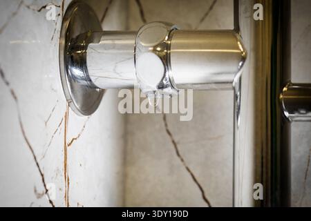 Single water drop falling from modern chrome plated towel warmer pipe, indicating plumbing leak or malfunction. Background of luxurious marble tiled b Stock Photo