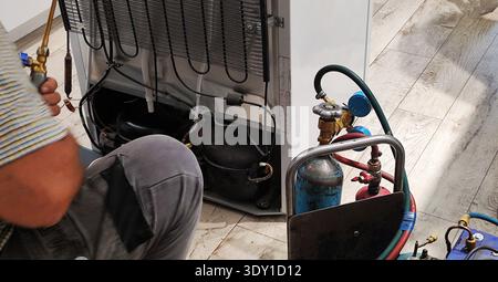 Person performing repair on back of domestic refrigerator. Technician hands working with torch, copper pipes. Refrigerant tanks, pressure gauges, hose Stock Photo
