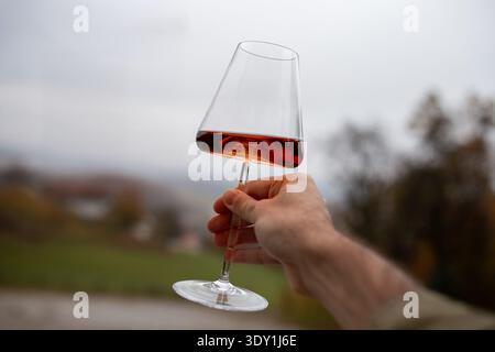 A hand holds an elegantly designed crystal wine glass, tilted to enhance the view of red dessert wine. The glass reflects a rich, amber-red hue, chara Stock Photo