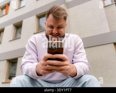 Young man sitting outdoors angry looking at his smartphone. Modern communication, social media, and digital lifestyle concept. Stock Photo