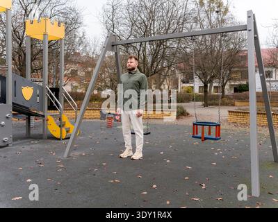 Bearded man standing under a swing set on a playground in a residential neighborhood. Calm thoughtful expression during an autumn day in a quiet urban Stock Photo