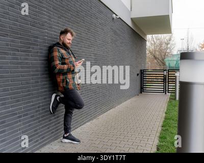 Relaxed young man leaning against wall and using smartphone. Mobile communication, modern technology and everyday urban lifestyle concept. Stock Photo