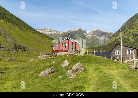 View of a vibrant red house nestled on a grassy hill beneath towering green mountains, with snow-capped peaks in the distance, Anestolen, Sogndal, Nor Stock Photo