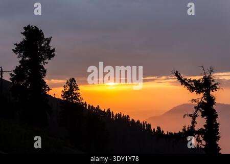 View of the sun sinking behind silhouetted trees on a mountain ridge with a warm glow illuminating the sky, Pir Chanasi, Azad Kashmir, Pakistan. Stock Photo