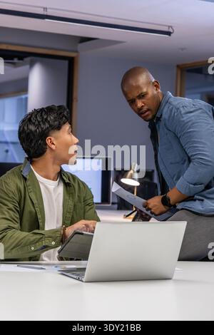 Diverse male coworkers collaborating at open office reviewing paperwork with laptop and tablet. Colleague, collaboration, discussion, planning, chart, Stock Photo