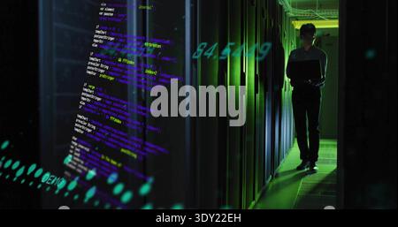 Standing Korean IT tech in dark uniform checking laptop at server aisle, projected code, copy space. Datacenter, rack, backlit, silhouette, overlay, n Stock Photo