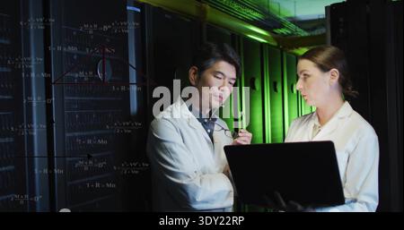 Analyzing two professionals in lab coats reviewing laptop in server room, with projected diagrams. Datacenter, collaboration, technicians, racks, equi Stock Photo
