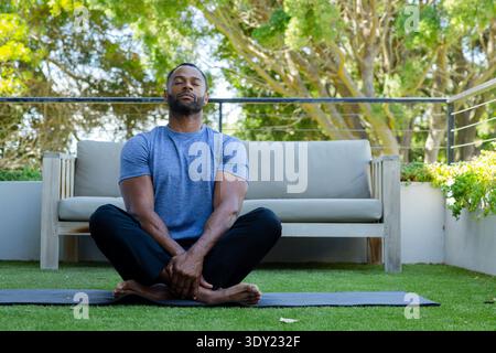 African American man meditating on black mat on patio wearing blue shirt by wooden sofa. Outdoor, terrace, turf, yoga, mindfulness, serenity, nature Stock Photo